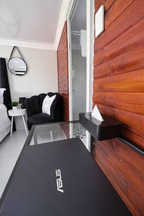 A modern hotel room with a glass-top table, a black ASUS laptop, a black sofa, a small side table with a lamp, and a wooden wall panel.
