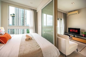 A hotel room with a large bed, a TV, a desk, and a view of high-rise buildings. - Sea View Balcony Condo | Gym + King Bed | 1min to Private Beach (Sattahip)