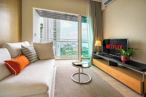 A modern hotel room with a sofa, a small table, a TV displaying Netflix, and a view of the city through large windows. - Sea View Balcony Condo | Gym + King Bed | 1min to Private Beach (Sattahip)