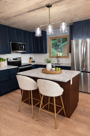 A modern kitchen with dark blue cabinets, stainless steel appliances, a central island with stools, and a wooden ceiling.