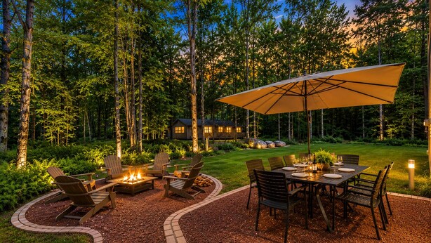 A patio with a fire pit, outdoor seating, and a table set for dining, surrounded by a wooded area and a house in the background. - Spacious 4-bedroom house in charming Arrowhead Lake w/ Fire Pit, Gameroom, Pool (pocono lake)
