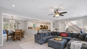 A spacious living room with a gray sectional sofa, a dining area with wooden chairs, and a kitchen in the background. - Spacious 4-bedroom house in charming Arrowhead Lake w/ Fire Pit, Gameroom, Pool (pocono lake)