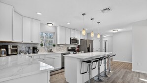 A modern kitchen with white cabinetry, a large island with bar stools, stainless steel appliances, and pendant lighting. - Spacious 4-bedroom house in charming Arrowhead Lake w/ Fire Pit, Gameroom, Pool (pocono lake)