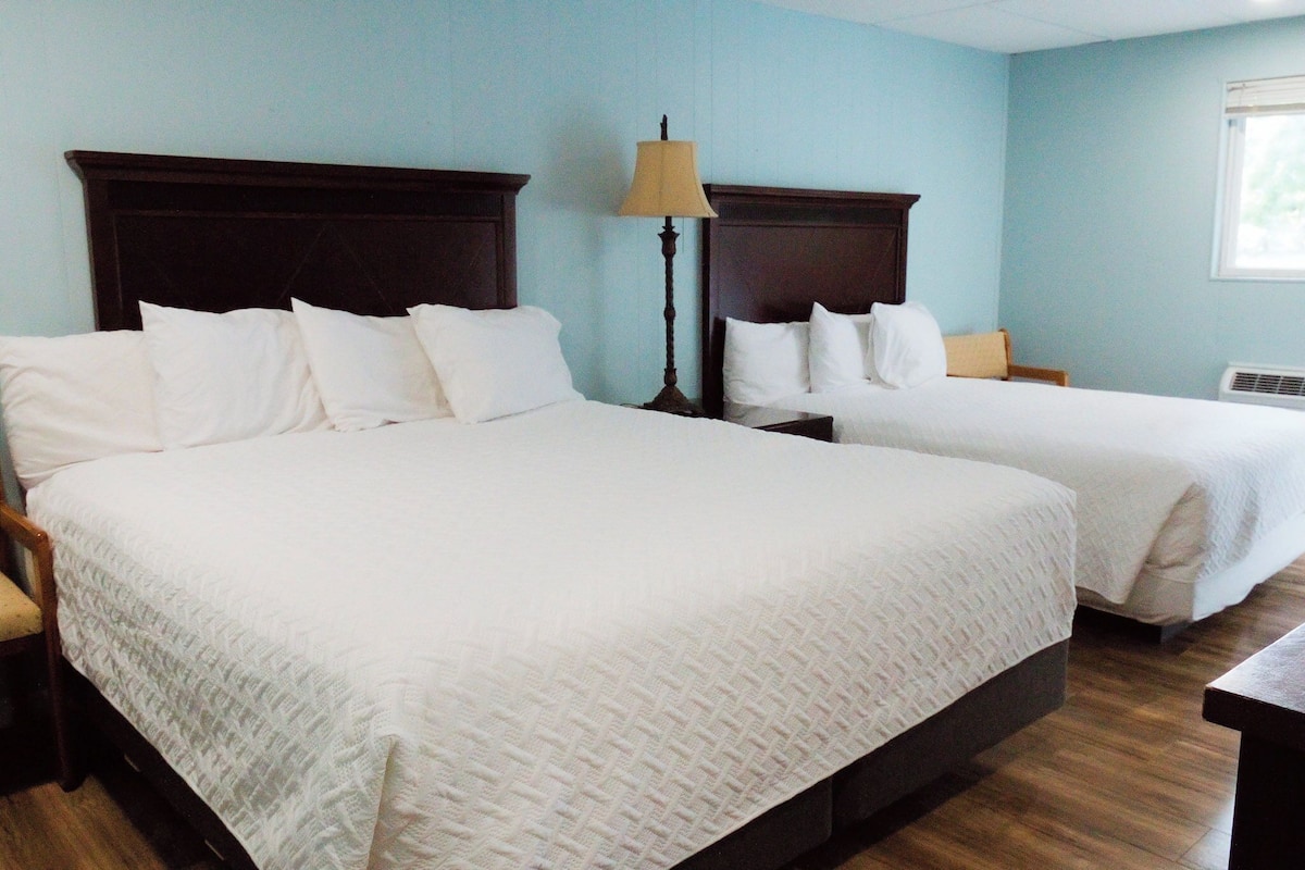 A hotel room with two beds, a nightstand, a lamp, and a window.