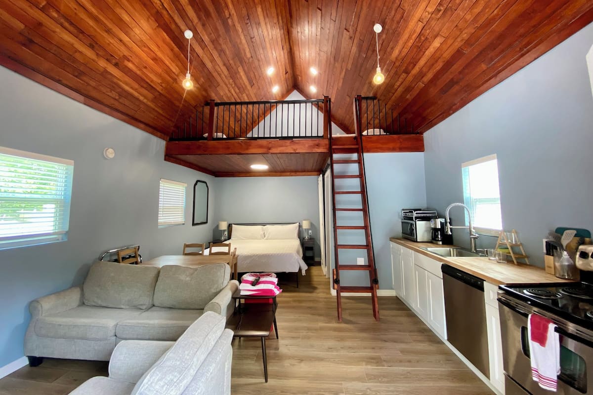 A loft-style room with a wooden ceiling, a loft bed, a kitchenette, a sofa, and a dining table.