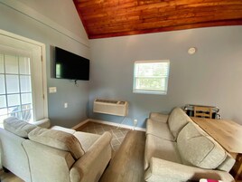 Una habitación con techo de madera, un televisor de pantalla plana montado en la pared, una ventana con persianas, un sofá y una pequeña mesa.