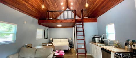 Una habitación estilo loft con techo de madera, una cama en alto, kitchenette, sofá y mesa de comedor.