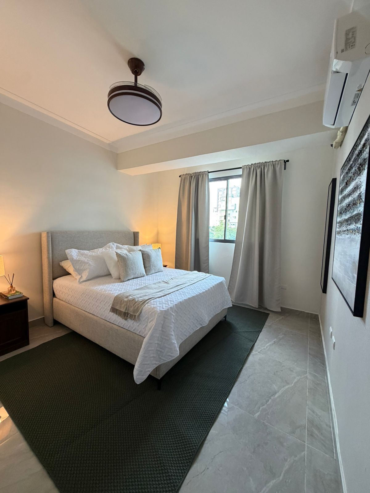 A bedroom with a bed, bedside lamps, a ceiling fan, a window with curtains, and a wall-mounted air conditioner.