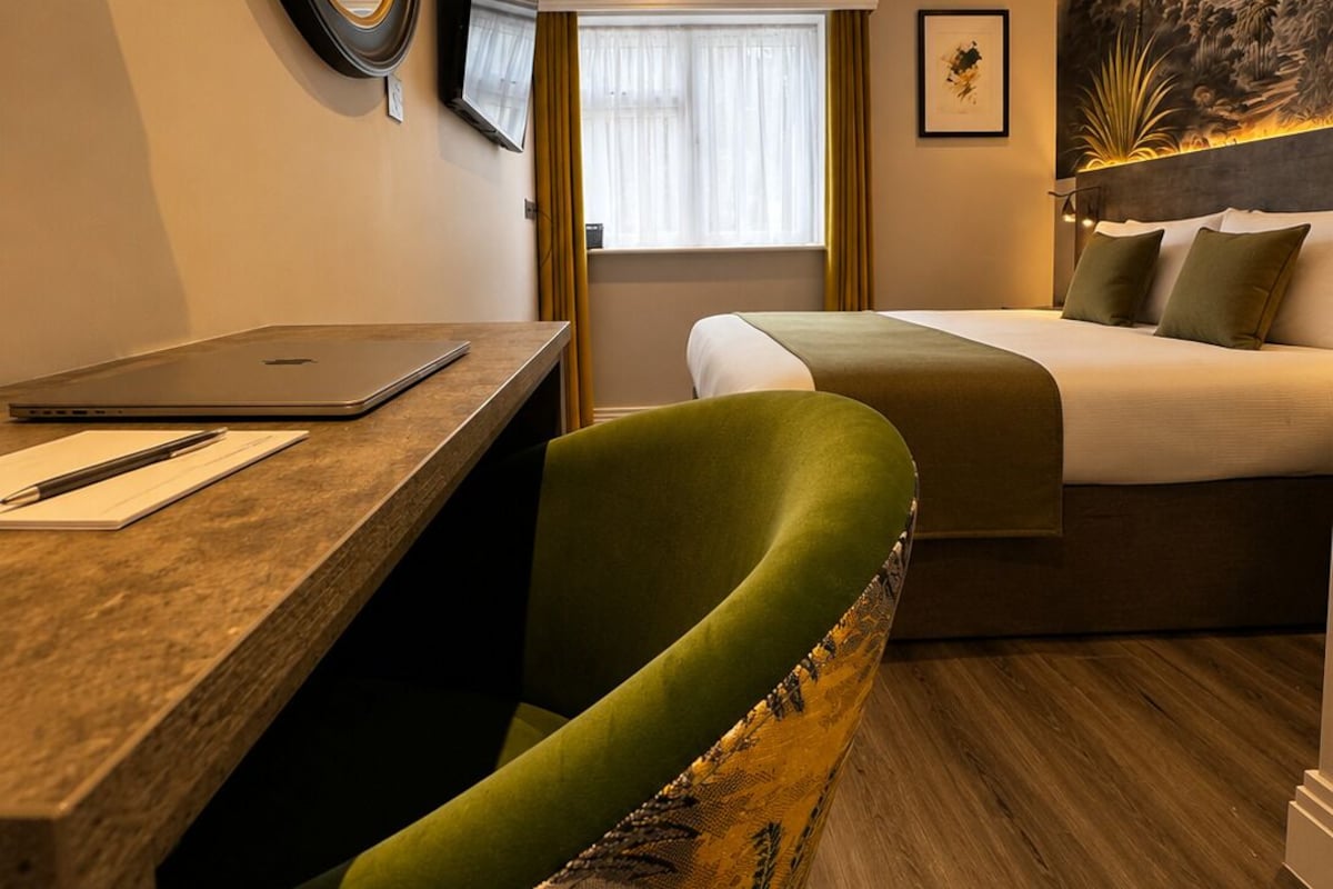 A hotel room with a desk, a green chair, a bed, a television, and a painting on the wall.