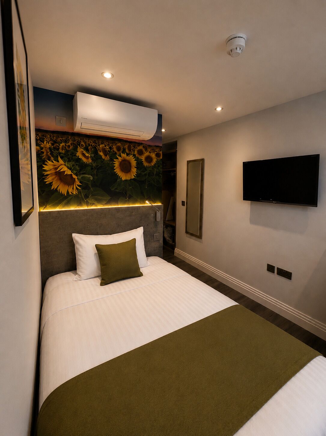 Quarto de hotel com cama, TV fixada na parede e ar condicionado, além de uma parede com um mural de girassóis.