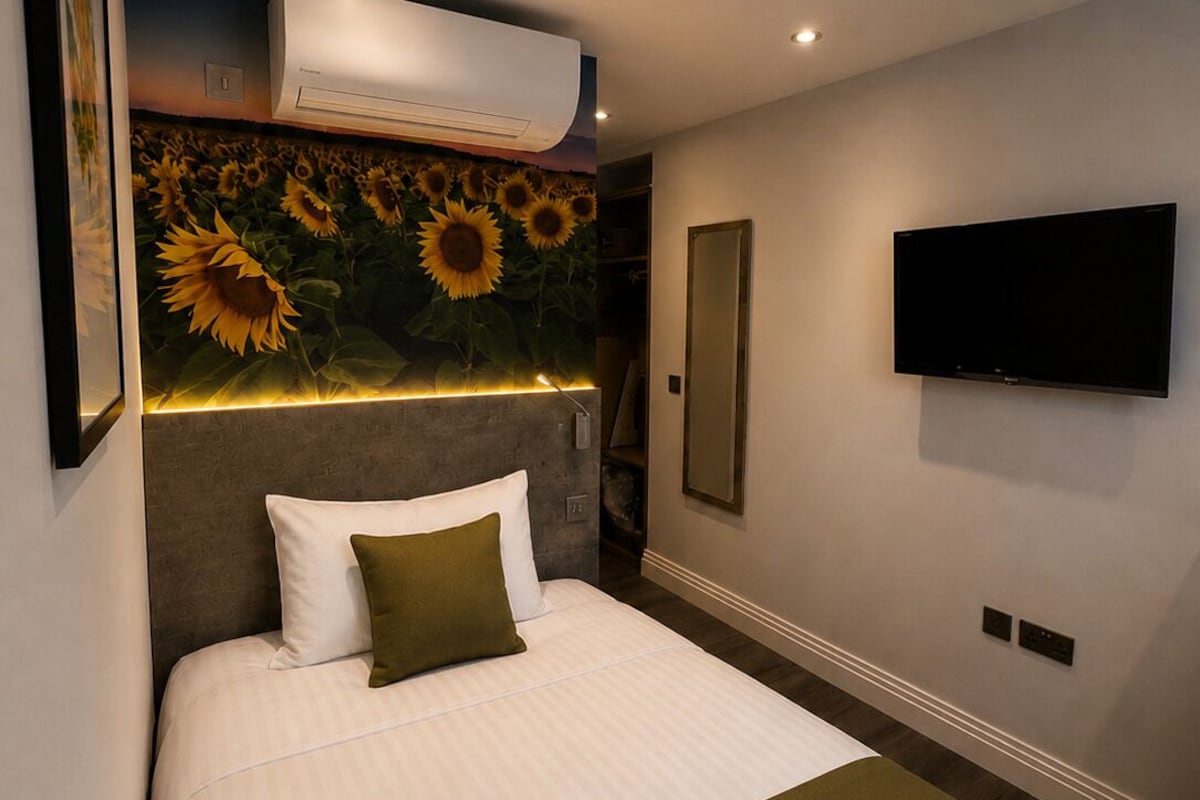 A hotel room with a bed, a mounted TV, an air conditioning unit, and a wall with a sunflower mural.