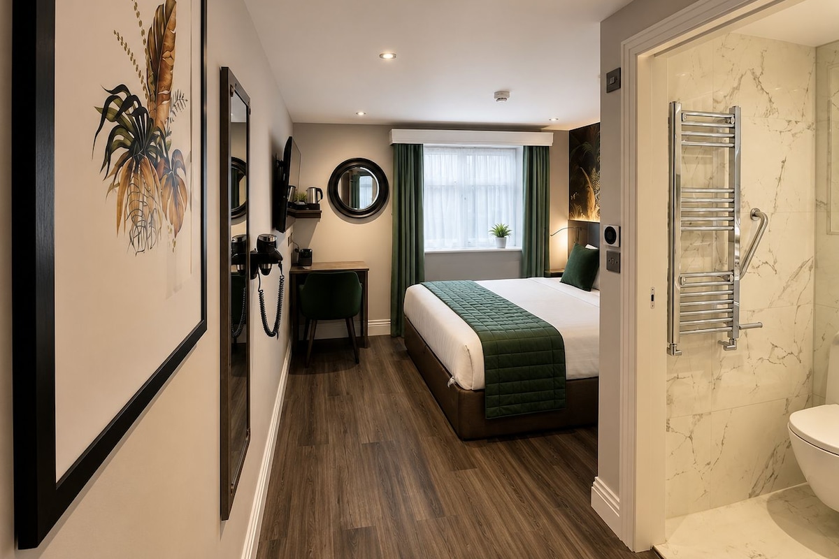 A hotel room with a bed, a desk, a mirror, a window with curtains, a bathroom with a toilet, and a shower area.