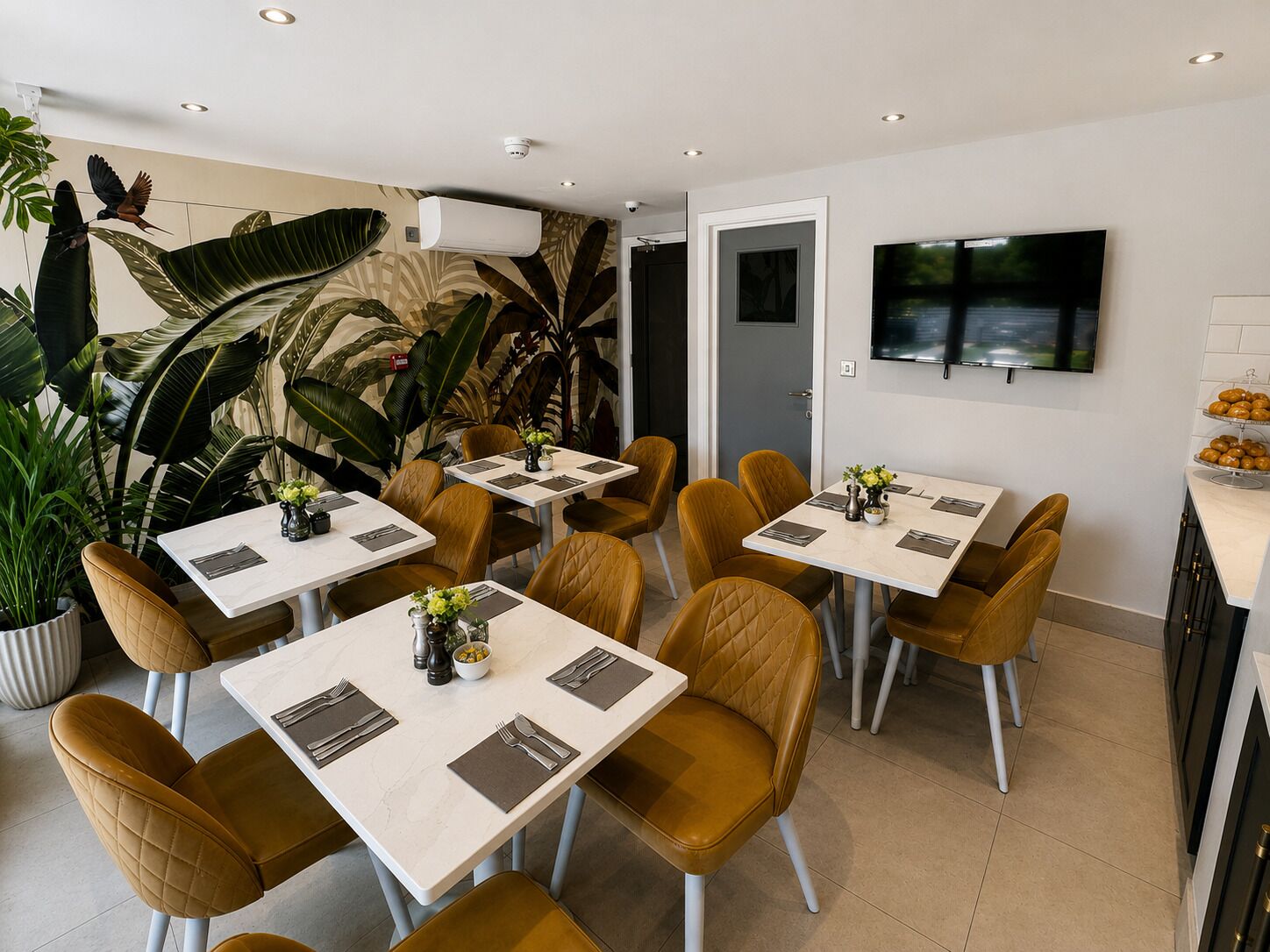 Uma área de refeições moderna com mesas, cadeiras, uma TV na parede e um mural de plantas.
