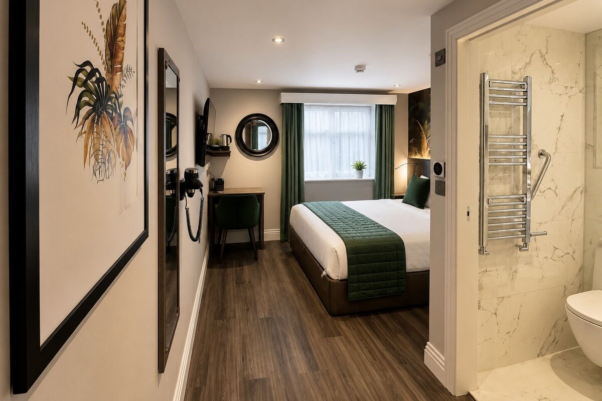 A hotel room with a bed, a desk, a mirror, a window with curtains, a bathroom with a toilet, and a shower area.