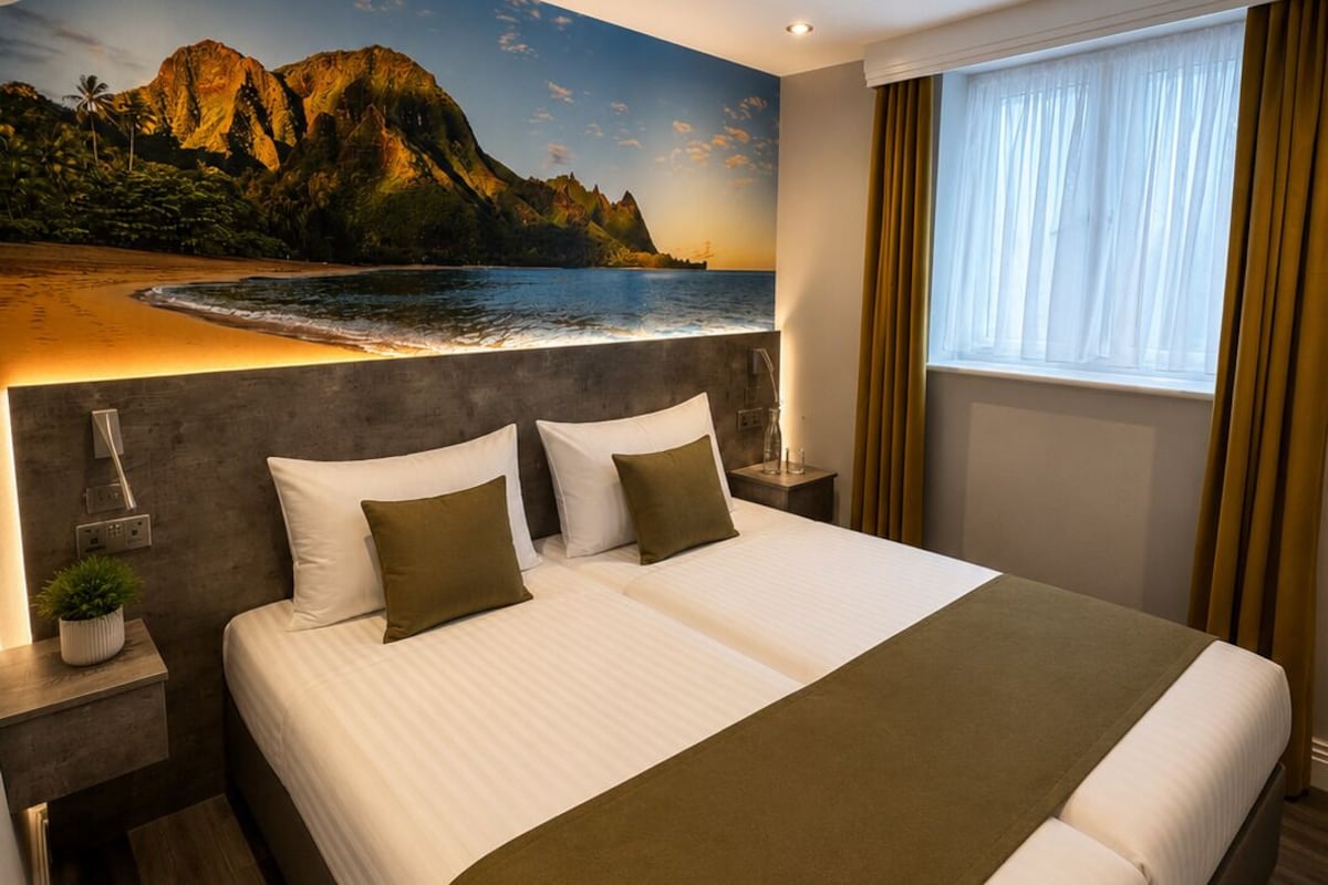 A hotel room with a large bed, a nightstand, a lamp, a window with curtains, and a wall mural of a beach scene.