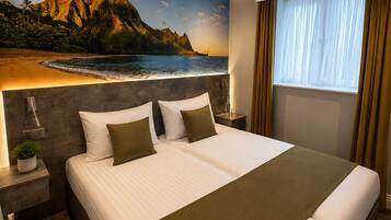 Quarto de hotel com uma cama grande, mesa de cabeceira, luminária, janela com cortinas e um mural na parede com uma paisagem de praia.