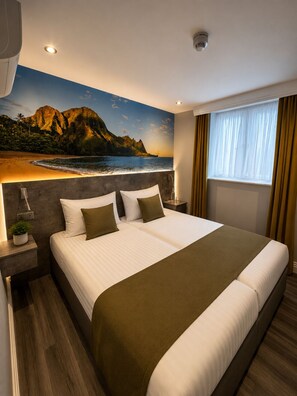 A hotel room with a large bed, a nightstand, a lamp, a window with curtains, and a wall mural of a beach scene. - Vivatar Heathrow Hotel (Hounslow)