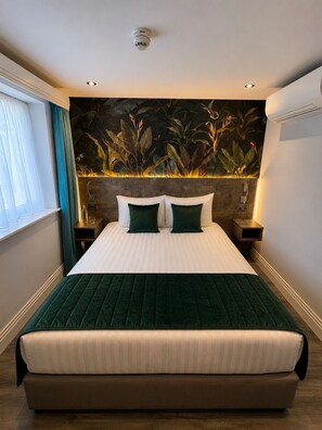 Habitación de hotel con una cama grande, una mesita de noche con lámpara, aire acondicionado instalado en la pared y una ventana con cortinas.