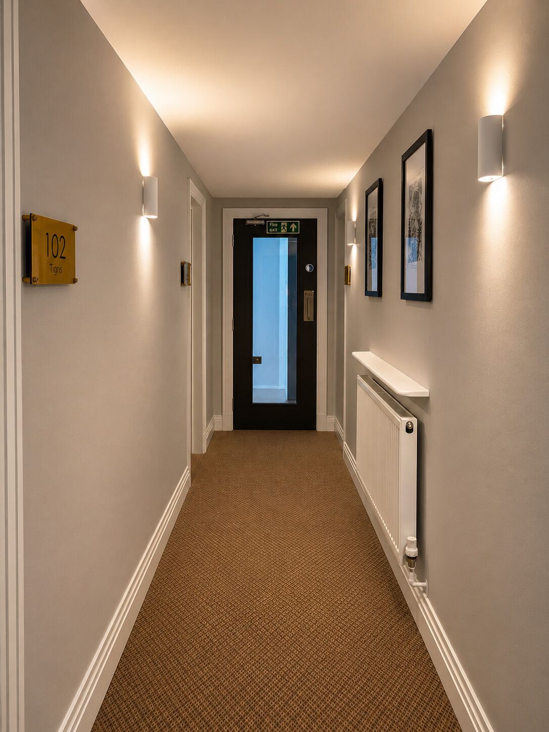 A hallway with a carpeted floor, a radiator, framed pictures on the wall, and a door with a fire exit sign.