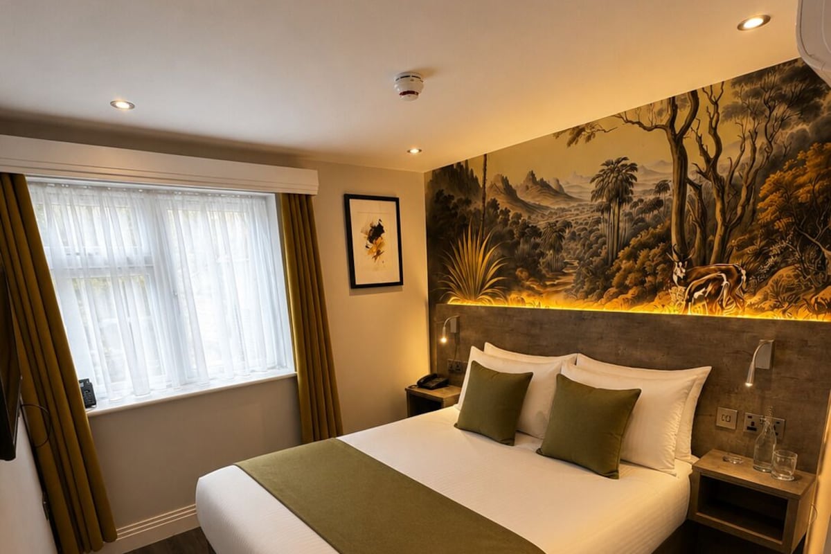 A hotel room with a large bed, a nightstand, a wall-mounted lamp, a painting, and a window with curtains.