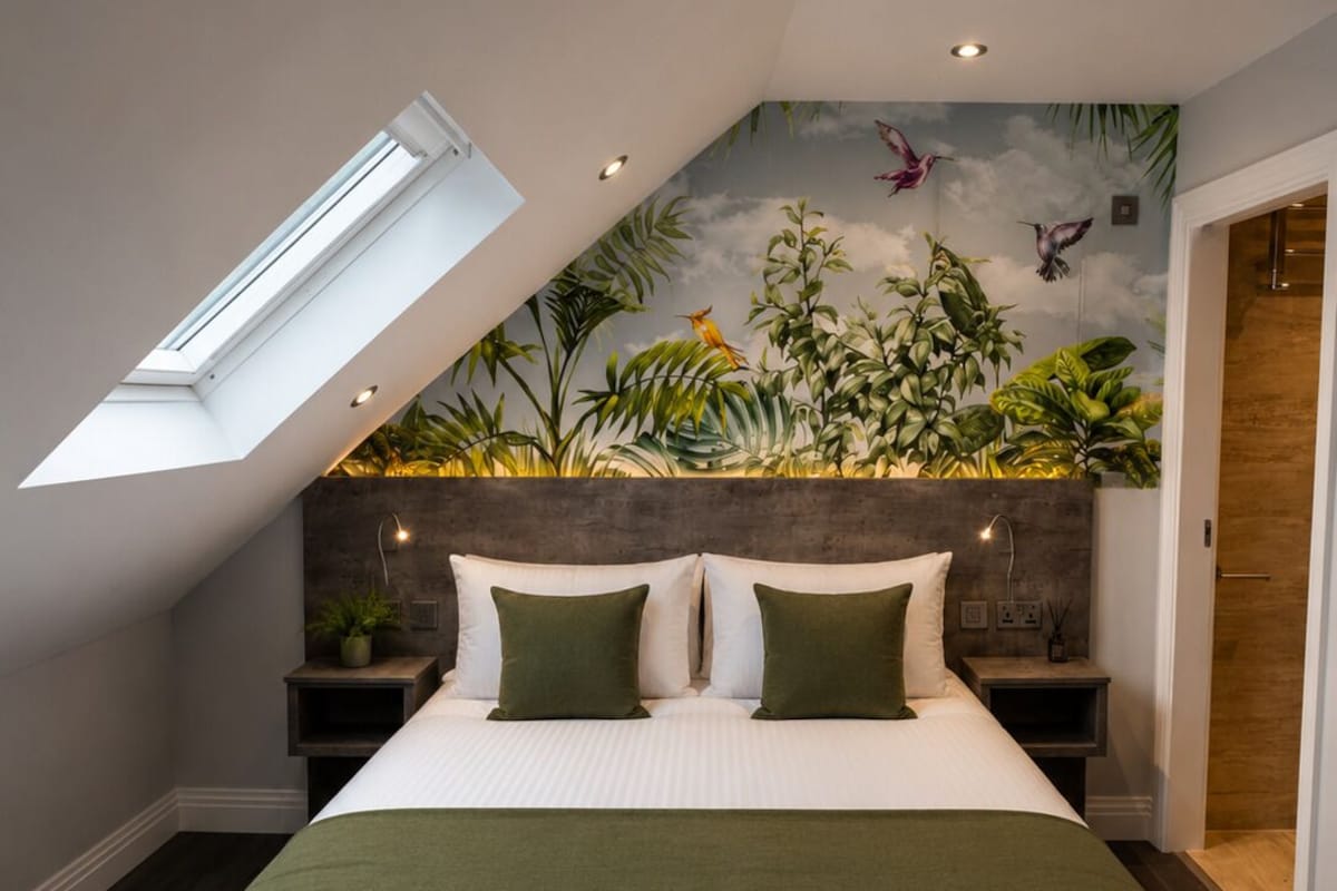 A bedroom with a skylight, a bed with green bedding, a mural of tropical plants, and a wooden headboard.