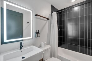 Uma casa de banho com uma pia branca, um espelho com luz, uma sanita, uma banheira com azulejos pretos e um separador de chuveiro.