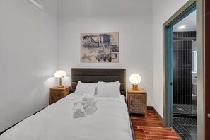 Uma cama bem arrumada com roupa de cama branca, duas luminárias de cabeceira, um quadro abstrato na parede e piso de madeira.