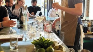 Een bar met een barman die drankjes klaarmaakt, een tafel met glazen en flessen, en een raam met uitzicht.