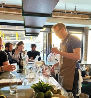 Uma área de bar com um bartender preparando bebidas, um menu e uma mesa com copos e garrafas.