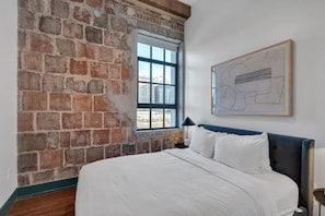 A bedroom with a brick wall, a large window, a bed with white bedding, and a framed abstract artwork on the wall. - 3 Bedrooms, French Quarter, Central Location, Pool, Walkable (New Orleans)