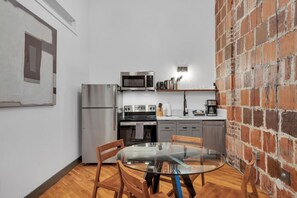 Une cuisine compacte équipée d’un réfrigérateur, d’un four à micro-ondes, d’un four et d’une machine à café. On y trouve une table en verre avec des chaises et un mur en briques.