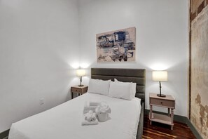 Un letto rifatto con lenzuola bianche, due comodini con lampade, una testiera e un'opera d'arte astratta appesa al muro.
