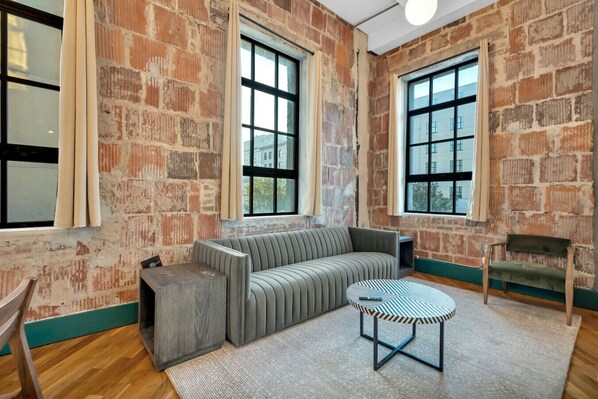 Quarto com paredes de tijolos aparentes, janelas amplas, sofá, mesa pequena e uma cadeira.