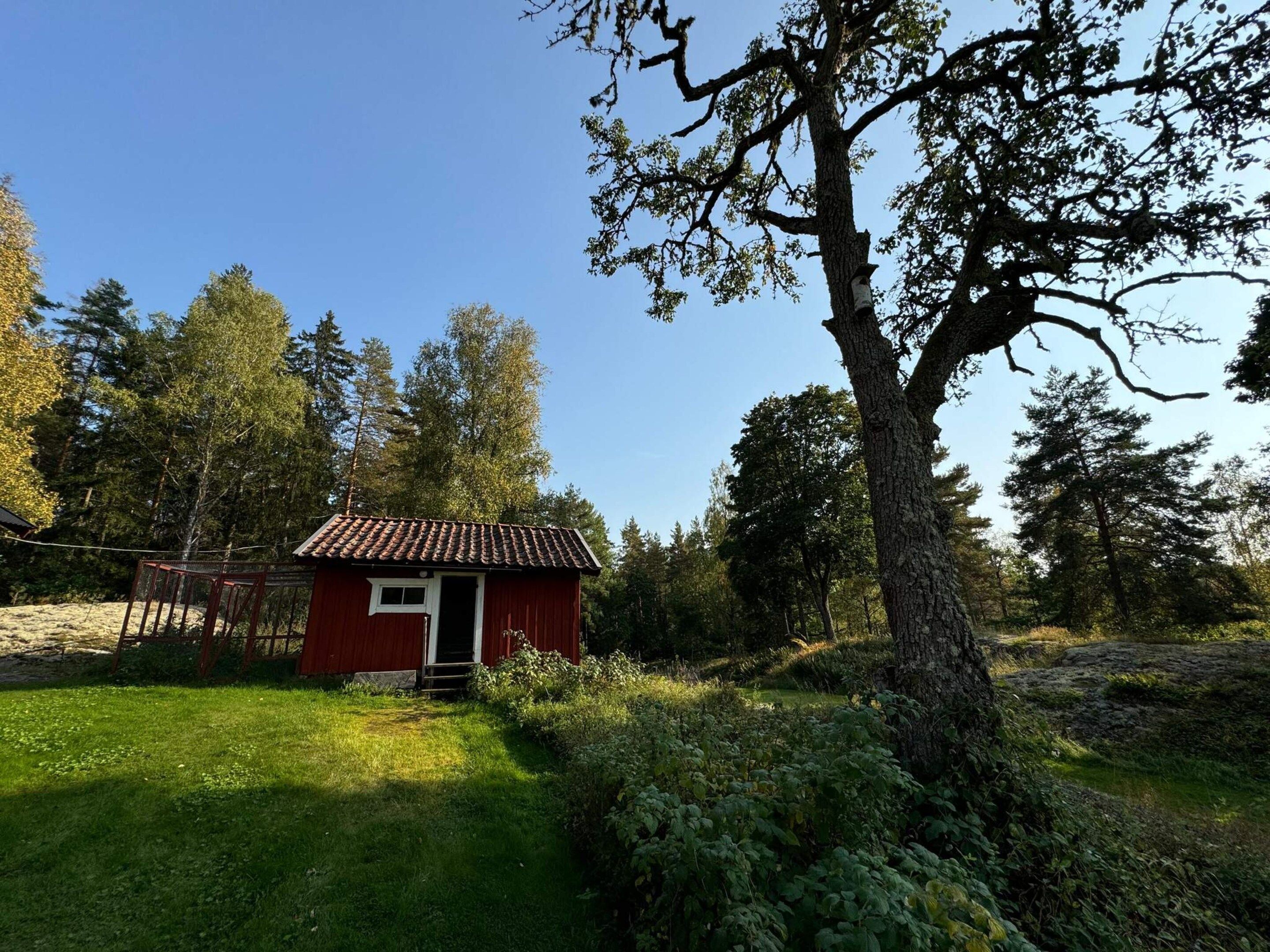 A red wooden cabin with a tiled roof, surrounded by a natural landscape with trees and a grassy area.