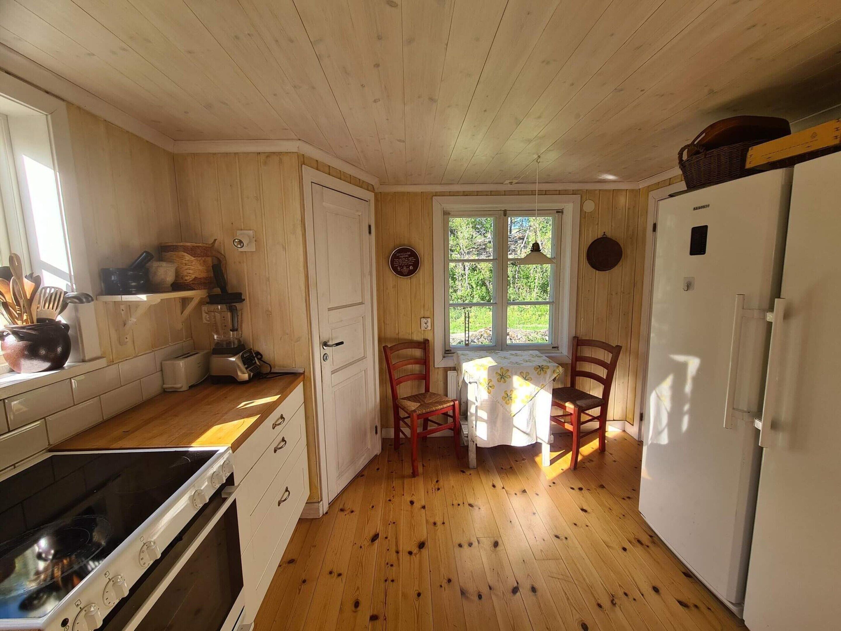 A kitchen with wooden walls, a dining table with chairs, a refrigerator, and a window with a view of greenery.