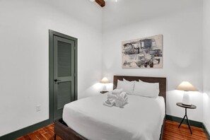 Quarto com uma cama, duas luminárias de cabeceira, um quadro, uma porta e piso de madeira.
