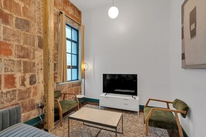 A room with a brick wall, a large window, a flat-screen TV, a white cabinet, and two green chairs.