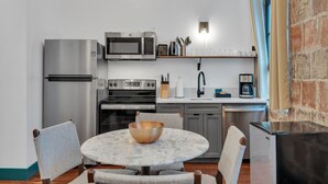 Cocina compacta con electrodomésticos de acero inoxidable, una mesa redonda con sillas y un muro de ladrillo.