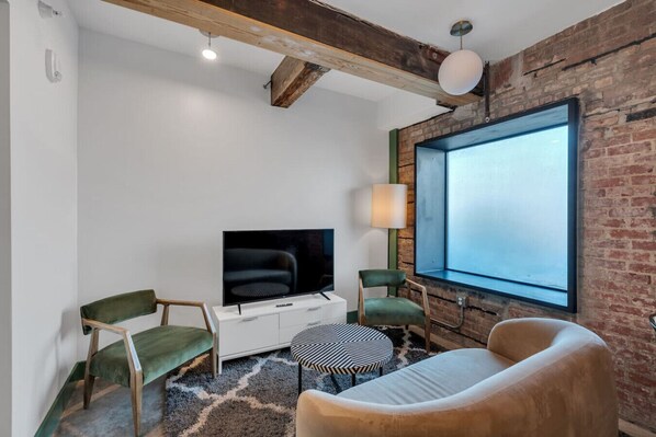 Un soggiorno moderno con una televisione a schermo piatto, due poltrone verdi, un divano bianco, un poggiapiedi decorato e un'ampia finestra con vista.
