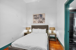 Una camera da letto moderna e accogliente con un letto singolo, due comodini con lampade, un quadro appeso e vista su una zona piastrellata attraverso una porta aperta.