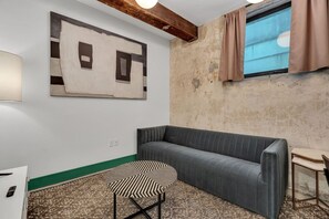 A room with a grey sofa, a patterned table, a painting on the wall, and a window with curtains.