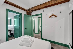 A hotel room with a bed, green-framed doors, a mirror, a hanging coat rack, and a white wall with a clock.
