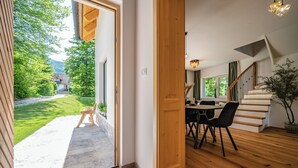 Un intérieur moderne avec un escalier en bois, un coin repas comprenant une table et des chaises, et une vue sur un paysage verdoyant par une porte ouverte.