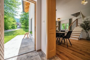 Ein modernes Interieur mit einer Holztreppe, einem Essbereich mit Tisch und Stühlen sowie Blick auf eine grüne Landschaft durch eine offene Tür.