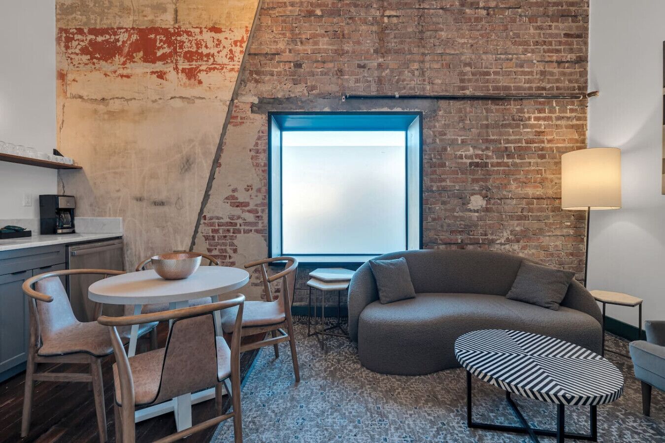 Un dormitorio estilo loft con pared de ladrillo, una mesa redonda de comedor con sillas, un sofá, un puf estampado y un ventanal grande.
