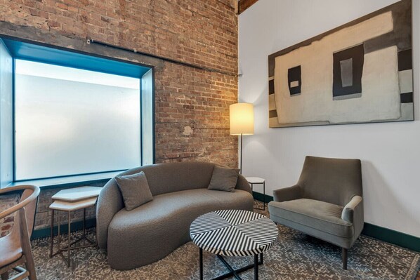 Una habitación con pared de ladrillo, una ventana grande, un sofá, una silla, una mesita y una lámpara de pie.