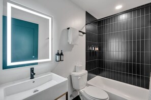 Un baño moderno con lavamanos, inodoro y bañera blancos, un espejo grande con puerta color turquesa y paredes con azulejos negros.