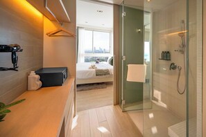 A modern hotel room with a bed, a shower, a small table with a radio, and a view of the city through large windows. - King Bed Sea View Condo | 2 Resort Pools | 1min to Private Beach (Sattahip)