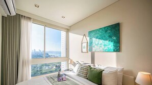 A hotel room with a bed, a desk, a laptop, a window with a city view, and a painting on the wall. - King Bed Sea View Condo | 2 Resort Pools | 1min to Private Beach (Sattahip)