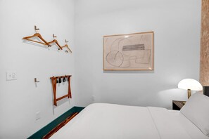 A hotel room with a bed, a wall-mounted coat rack, a framed abstract painting, and a bedside lamp.